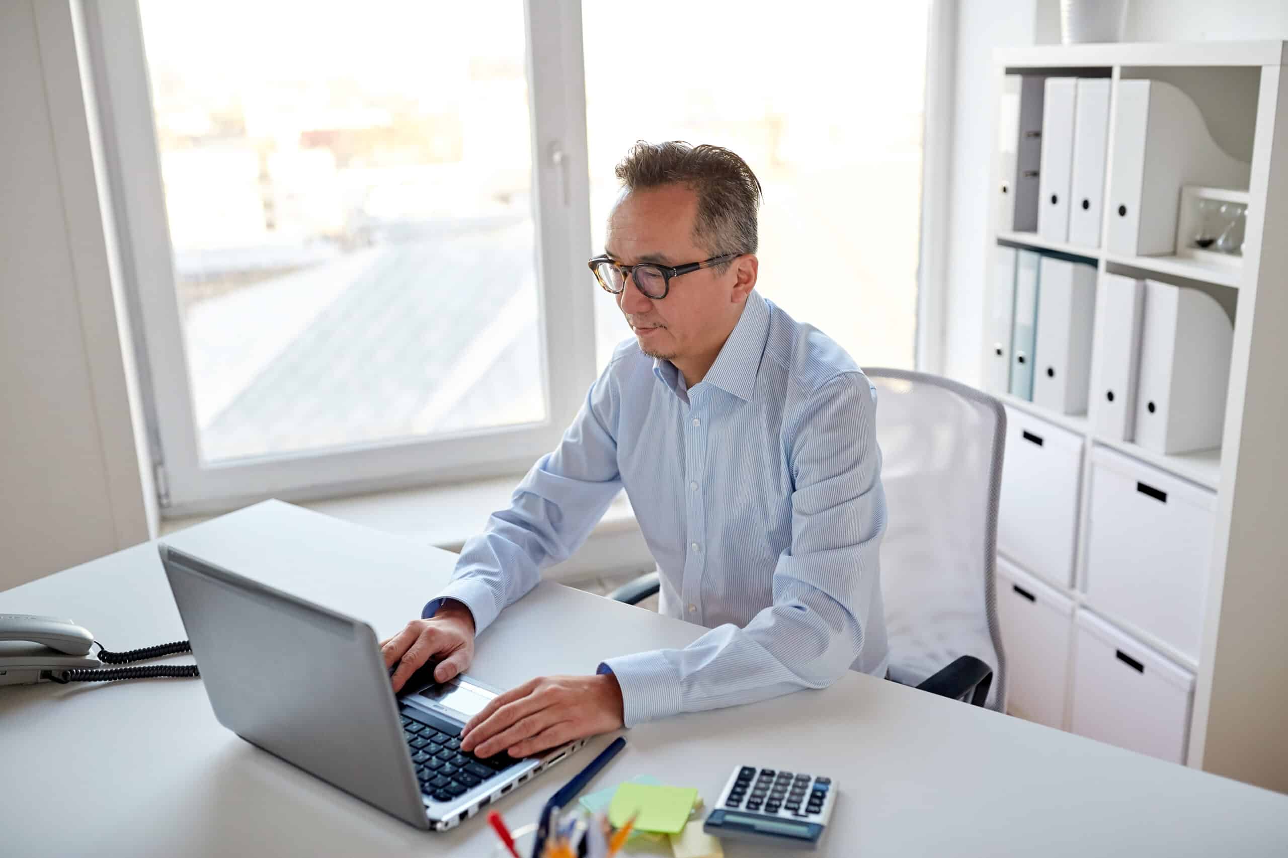 business, people and technology concept - businessman in eyeglasses with laptop computer office